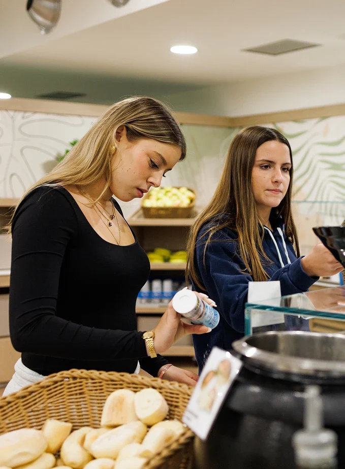 Nella mensa scolastica in primo piano si vede un cesto di piccoli panini, quindi una studentessa bionda dai capelli lunghi che sparge del sale da un barattolo, un’altra porge la mano destra