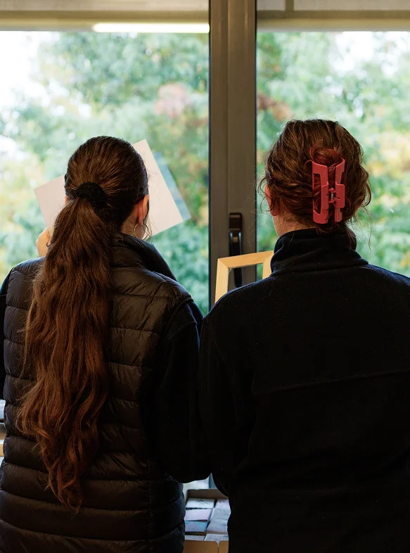 Due studentesse del club annuario scolastico dell’IST, viste di spalle, sono in piedi davanti a una finestra. La ragazza di destra porta i capelli raccolti con una pinza rossa, quella di sinistra li ha sciolti