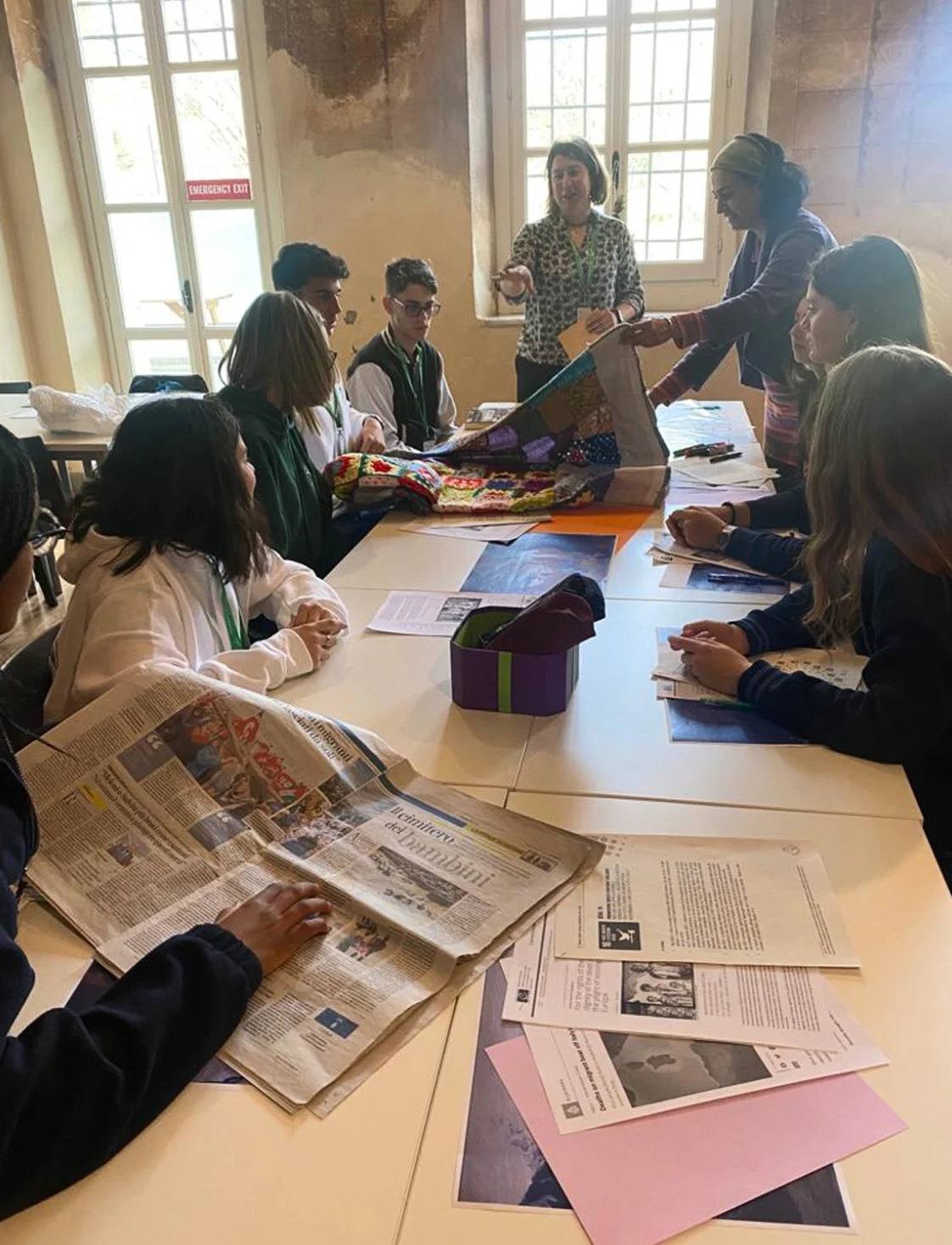 Riunione di uno dei club scolastici dell’IST. Gli studenti sono seduti a un lungo tavolo di lavoro bianco, dispongono di giornali e fotocopie ed esaminano una coperta patchwork all’uncinetto