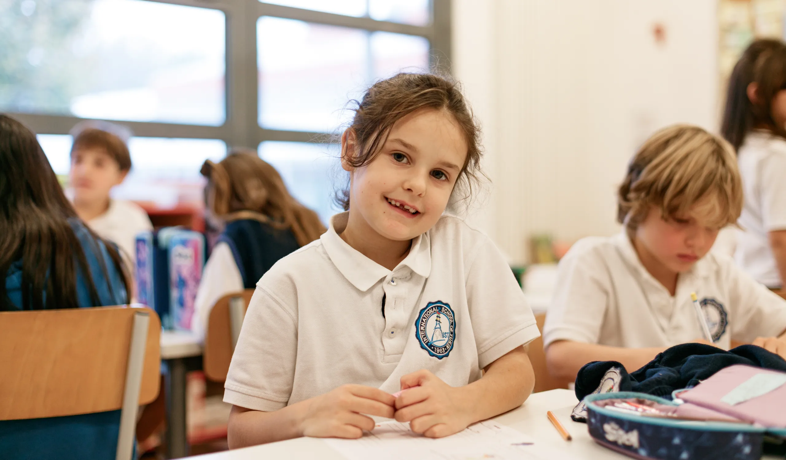 Scolari di IST al lavoro: in primo piano una bambina con i capelli castani raccolti e la polo di IST guarda l’osservatore e sorride, mostrando gli incisivi permanenti in crescita