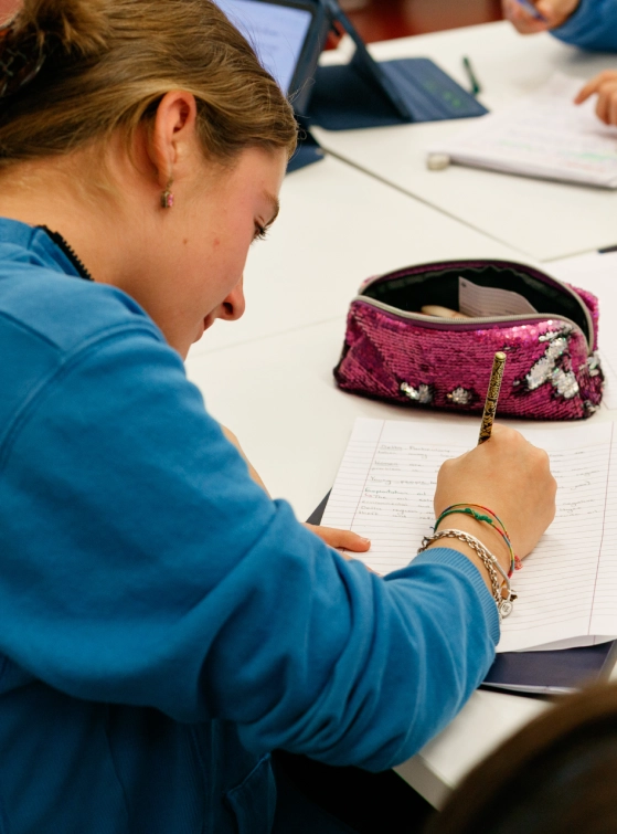 Una giovane donna con una felpa azzurra, seduta a una scrivania bianca, scrive con la mano destra su un foglio protocollo a righe. Davanti a lei si trova un astuccio rosa con disegni bianchi