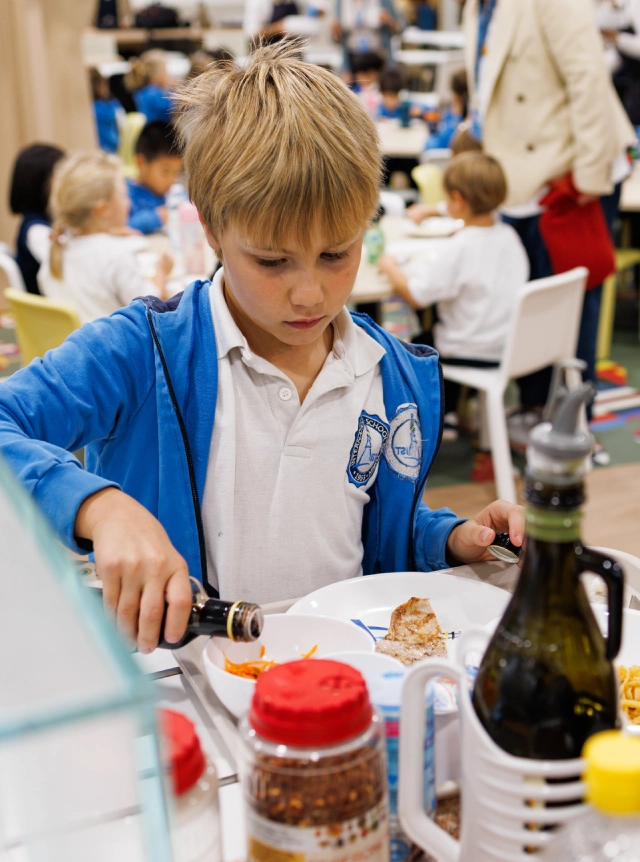 Un ragazzo biondo con una felpa azzurra dell’IST, a un tavolo della mensa scolastica, versa con la destra del condimento da una bottiglia in una scodella con insalata di carote