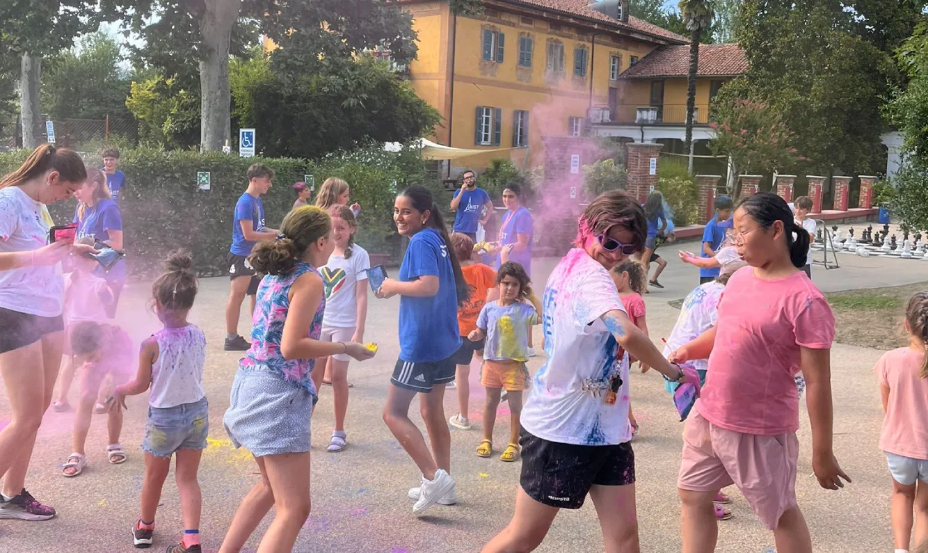 Ragazzi e bambini in tenuta estiva giocano con polveri colorate nel campo scuola estivo di IST. Sullo sfondo il corpo principale della scuola, edificio arancione con tetto in coppi rossi