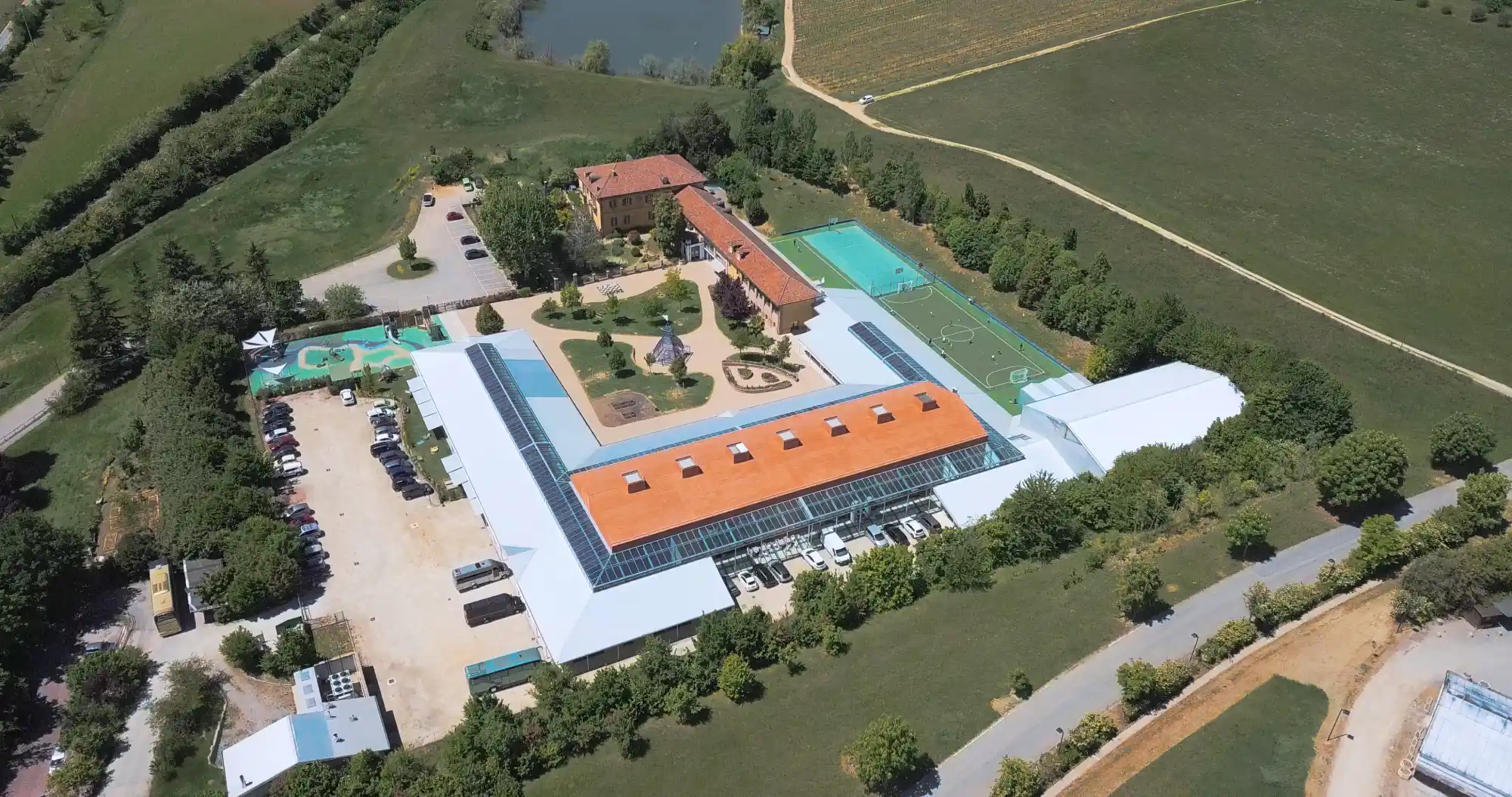 Visione aerea di IST, tra campi coltivati, con ampi parcheggi, campus e area giochi interni, palestra coperta e due campi sportivi