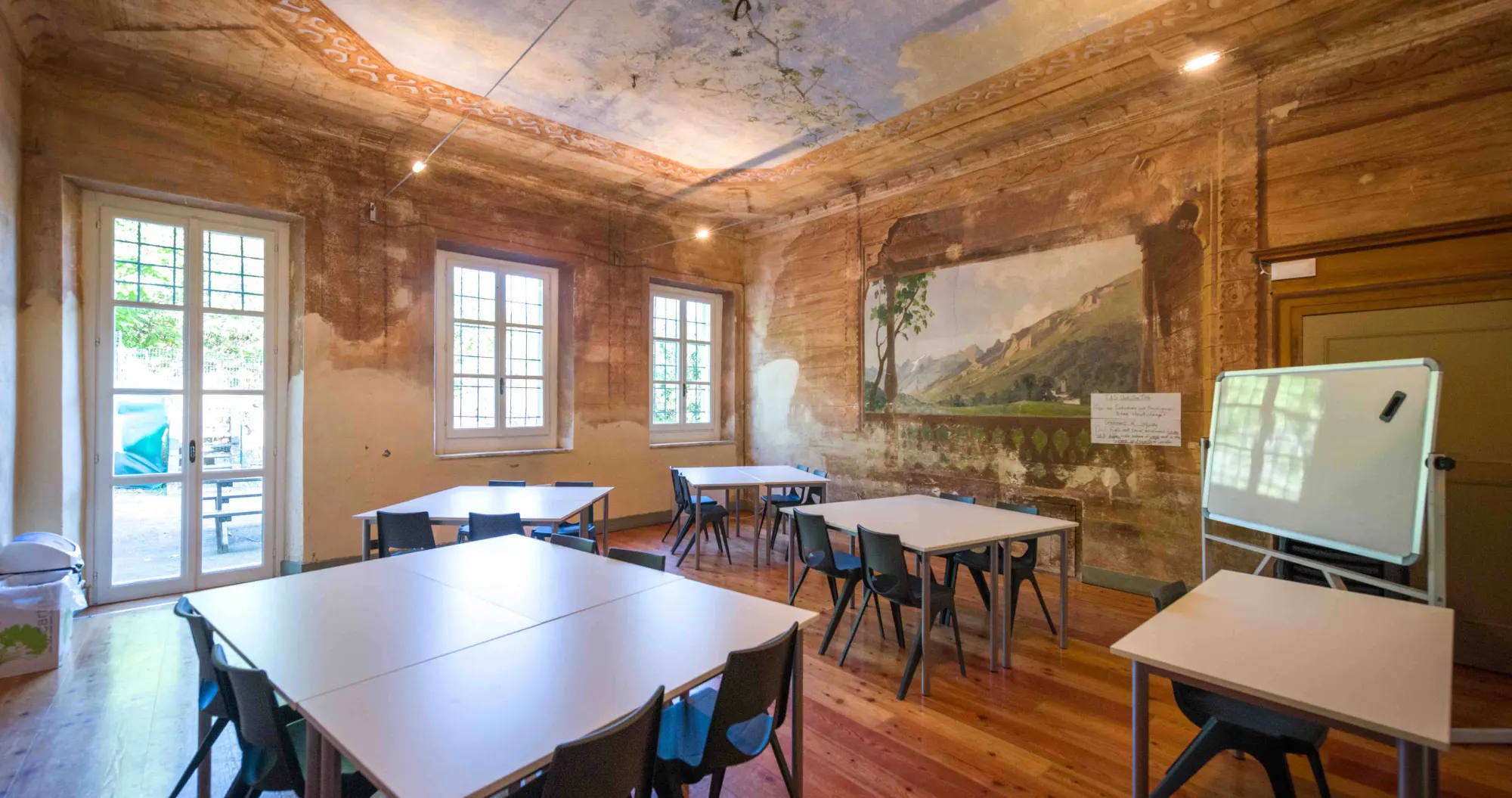 Salone con pavimento in legno, pareti e soffitto affrescati e restaurati. Cinque tavoli da lavoro bianchi con sedie in plastica scura. A destra una lavagna bianca
