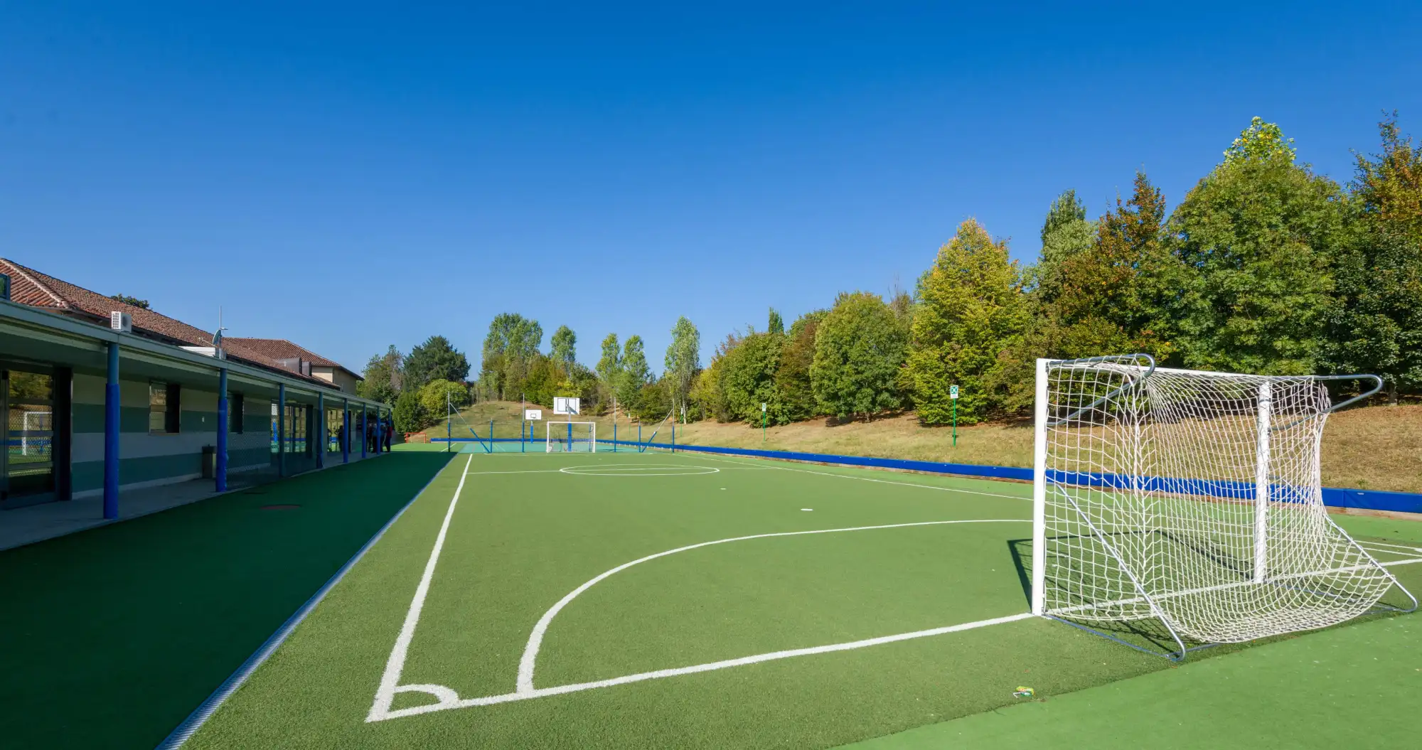 Campo da calcio in erba sintetica verde con porte bianche. A destra zona verde fitta di alberi, a sinistra il porticato dell’IST campus torino