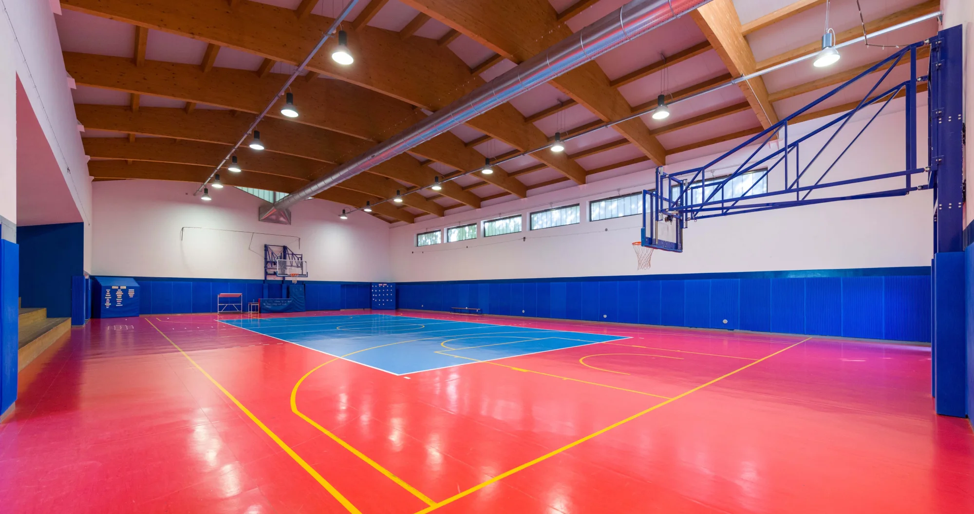 Interno di una grande palestra con capriate in legno e potenti luci sospese. Il pavimento rosso e azzurro corrisponde a un campo da basket