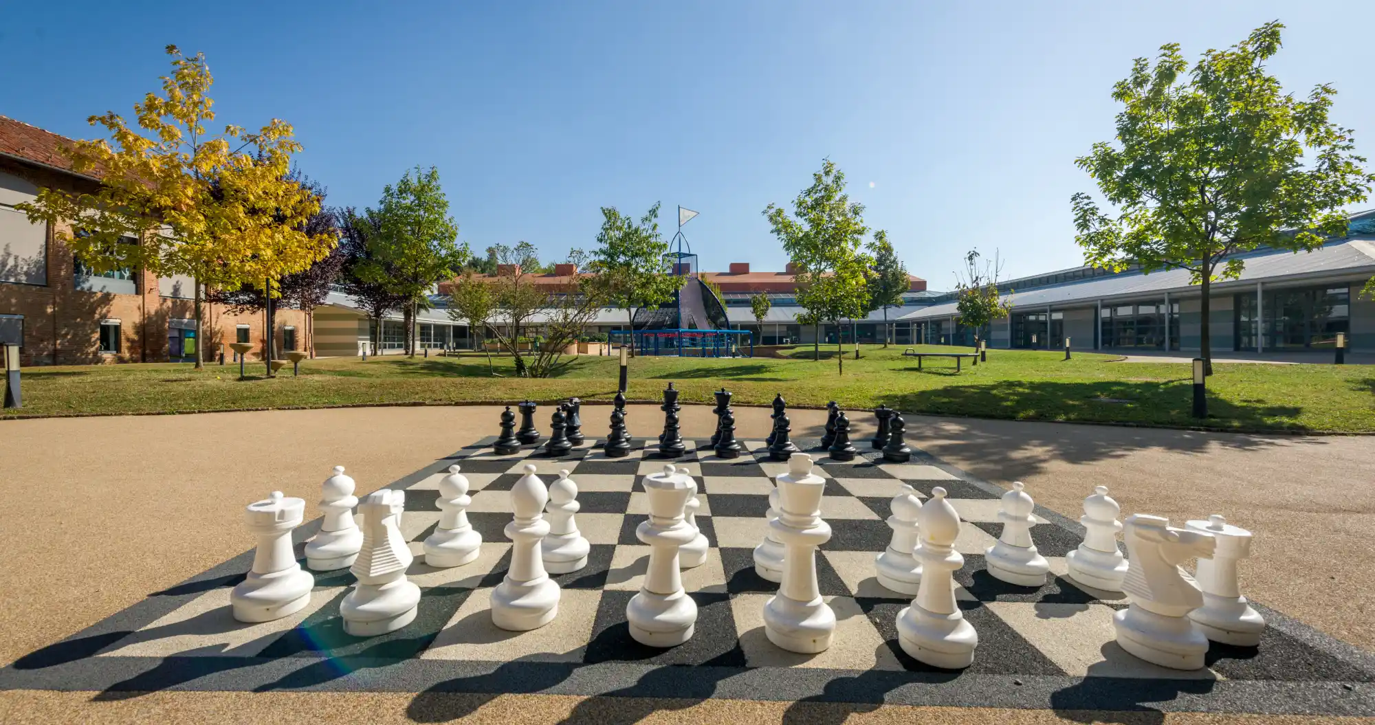 Scacchiera gigante dipinta sul terreno: i pezzi bianchi (in primo piano) e neri sono in posizione di gioco. Sullo sfondo il parco alberato di IST campus torino