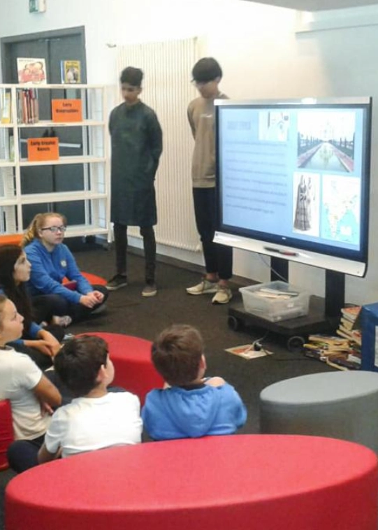 In un’aula scolastica sei bambini seduti a terra sulla moquette marrone tra pouf rossi e grigi seguono una presentazione di slides da parte di due ragazzi