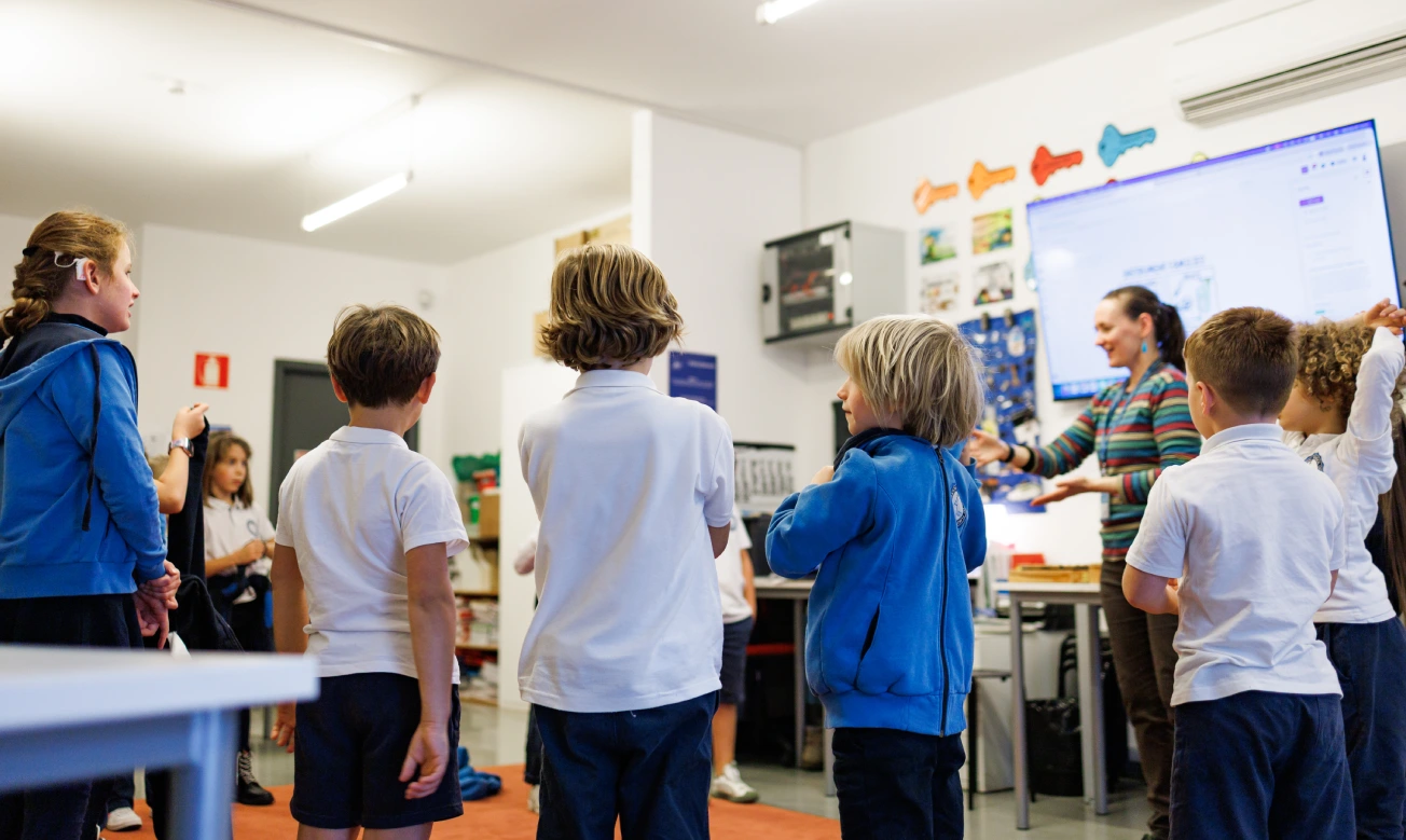 Insieme di scolari con la divisa azzurra, bianca e nera dell’IST durante una lezione da parte di un’insegnante con coda di cavallo castana e golfino a righe orizzontali multicolori