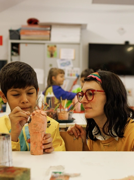 Un ragazzo dai capelli neri corti rifinisce una piccola scultura cilindrica in argilla, affiancato dall’insegnante con i capelli neri lisci a media lunghezza e con occhiali dalla montatura rossa