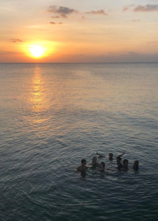 Un gruppo di sette giovani nuota nel mare calmo nel corso di un viaggio di scambi internazionali, il sole all’orizzonte sta tramontando, nel cielo poche nubi sparse